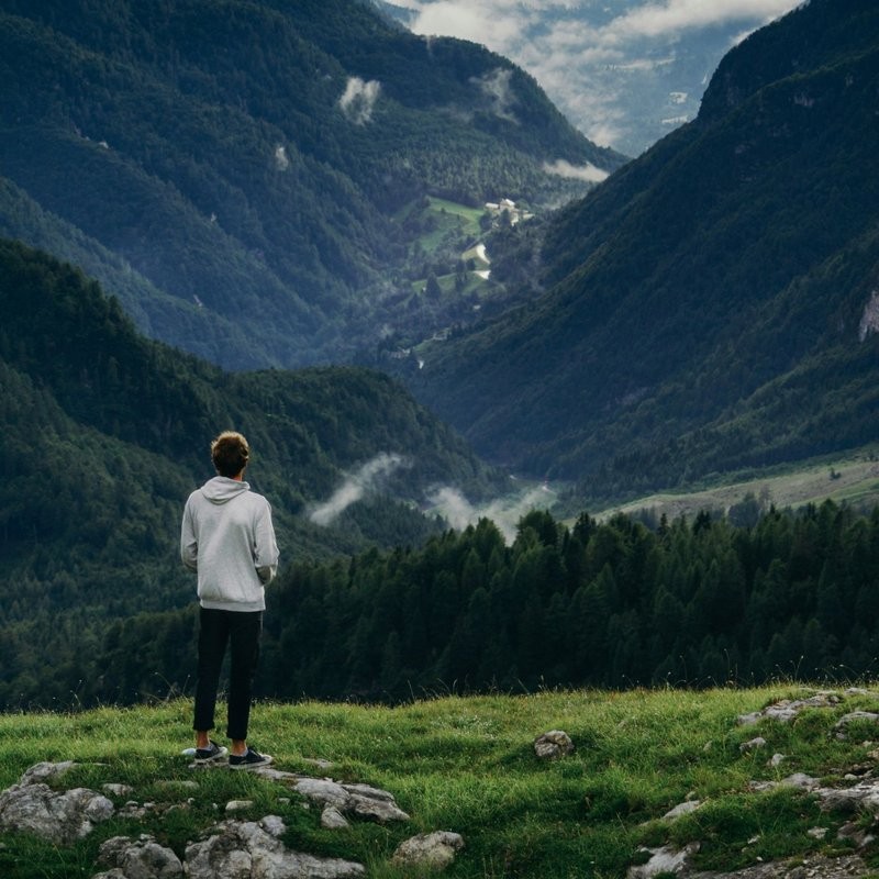 A person stands on a grassy ridge, looking out over a deep valley and the long, steep route ahead through cloud-filled mountains