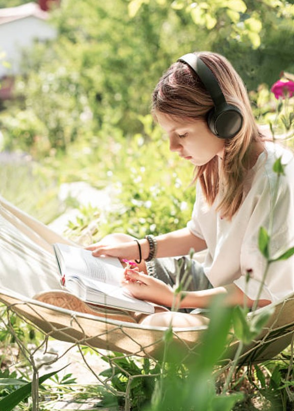 A teenager reading and highlighting a book in a garden hammock, wearing headphones.